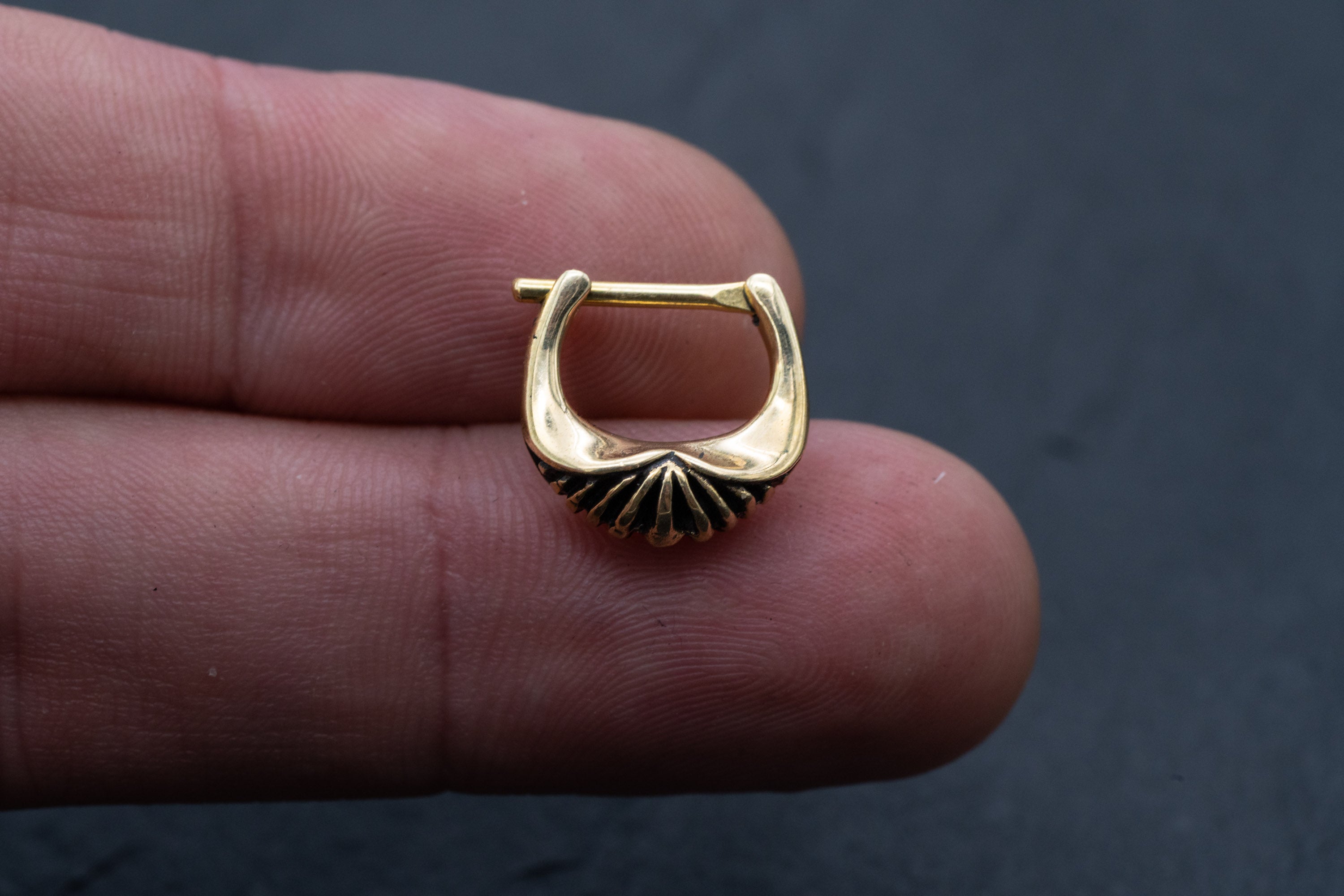 A person's fingers holding the DIARA Biomechanical Clicker Hoop Septum Nose Ring by Kaya Movement against a dark background. The unisex, 16-gauge gold nose ring features a ridged design pattern at the bottom.